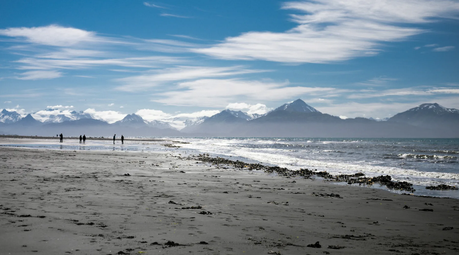 A wide beach with a few people walking, snowy mountains across the inlet